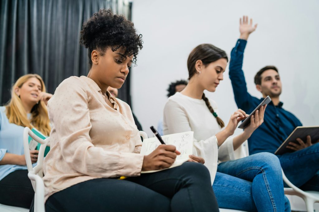 Attendees participating in a professional workshop, with one person taking notes, another using a tablet, and a participant raising a hand during a classroom discussion.